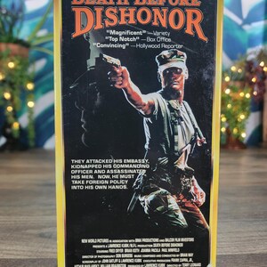 May include: A VHS tape case for the film Death Before Dishonor, featuring a soldier in camouflage holding a handgun. The cover includes the title in orange text, critical reviews, a plot summary, and production credits printed in white on black.