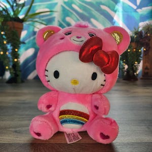 Pode incluir: Um brinquedo de pelúcia rosa com um design Hello Kitty com um capuz de urso e um laço vermelho. O brinquedo tem um design de arco-íris na barriga e detalhes dourados nas orelhas. A pelúcia está em uma superfície de madeira.