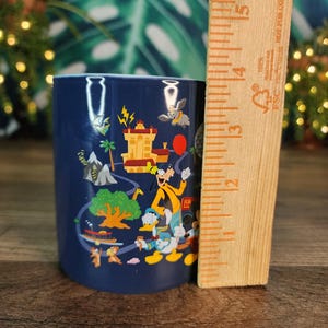 May include: A navy blue ceramic mug featuring colorful cartoon characters and scenes. The mug is approximately 4 inches tall and has a cylindrical shape. A wooden ruler is placed next to the mug for scale.