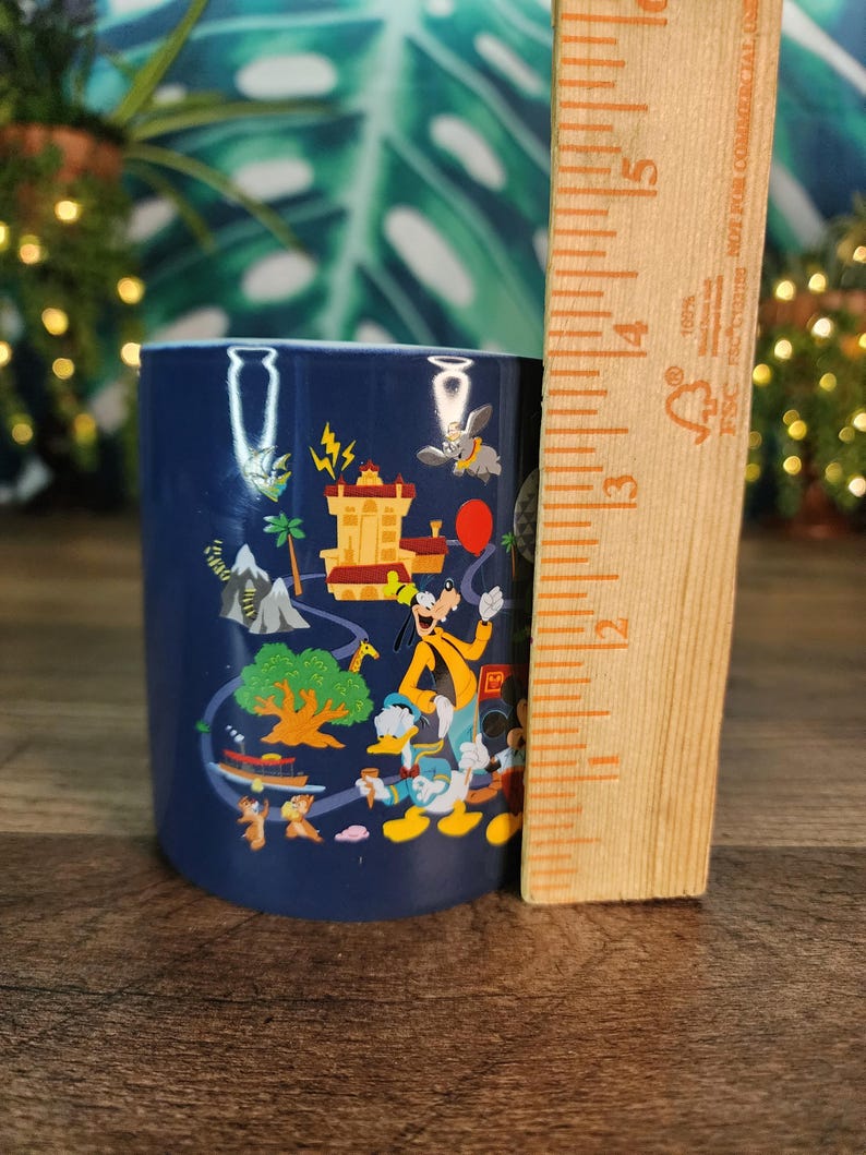 May include: A navy blue ceramic mug featuring colorful cartoon characters and scenes. The mug is approximately 4 inches tall and has a cylindrical shape. A wooden ruler is placed next to the mug for scale.