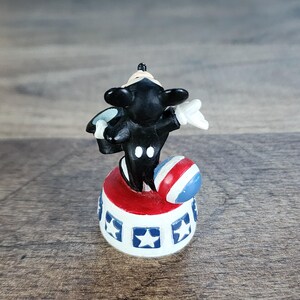May include: A small figurine of a black and white cartoon character balancing a red, white, and blue ball. The character is standing on a red and white platform with blue star accents. The figurine is a collectible item.