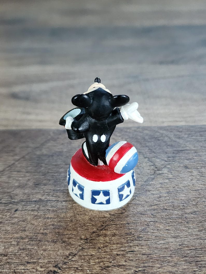 May include: A small figurine of a black and white cartoon character balancing a red, white, and blue ball. The character is standing on a red and white platform with blue star accents. The figurine is a collectible item.