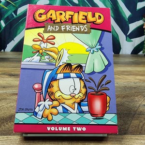 Puede incluir: Caja de DVD de Garfield y sus amigos, Volumen Dos. La portada presenta a Garfield en pijama sosteniendo una taza roja de café. El título es naranja y el fondo es una escena de dibujos animados colorida.