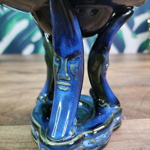 May include: A ceramic oil burner featuring a glossy dark blue and black glaze. The structure is supported by three curved legs, each sculpted with a detailed human face. The top section is a wide, circular bowl, and the base is a triangular platform.