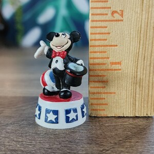 May include: A miniature Mickey Mouse figurine in a magician's outfit, holding a top hat. The figure stands on a red, white, and blue circus-style platform, with a ruler next to it. The figurine is approximately 1 inch tall.