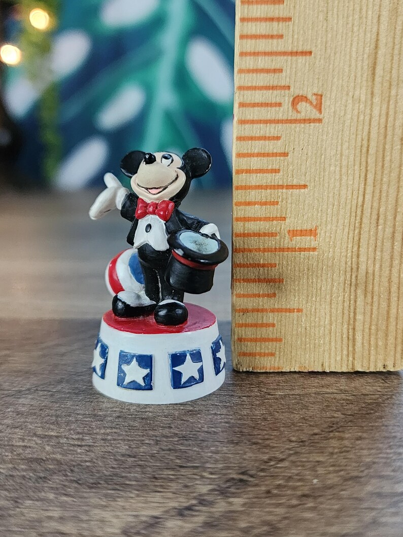 May include: A miniature Mickey Mouse figurine in a magician's outfit, holding a top hat. The figure stands on a red, white, and blue circus-style platform, with a ruler next to it. The figurine is approximately 1 inch tall.