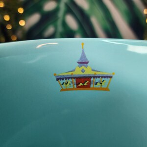 May include: A light blue ceramic bowl with a carousel design. The carousel has a yellow roof, purple trim, and red doors. The carousel has horses and riders. The background is blurred with green leaves and golden bokeh lights.