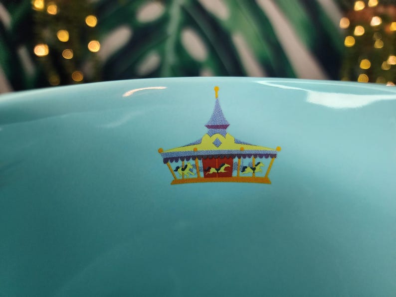 May include: A light blue ceramic bowl with a carousel design. The carousel has a yellow roof, purple trim, and red doors. The carousel has horses and riders. The background is blurred with green leaves and golden bokeh lights.