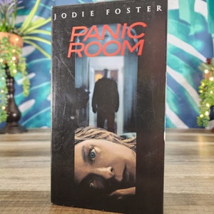 Puede incluir: Una cinta VHS negra de la película "Panic Room" con Jodie Foster. La portada muestra el título en rojo, una figura sombría en una puerta y un primer plano del rostro de una mujer.