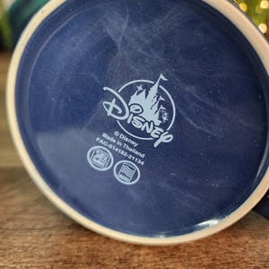 May include: A navy blue ceramic mug base with a white rim. The Disney logo is centered, featuring a castle silhouette. The text "Made in Thailand" and other details are visible. The mug sits on a wooden surface.