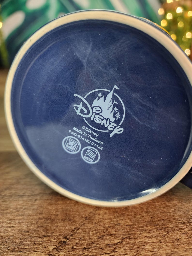 May include: A navy blue ceramic mug base with a white rim. The Disney logo is centered, featuring a castle silhouette. The text "Made in Thailand" and other details are visible. The mug sits on a wooden surface.