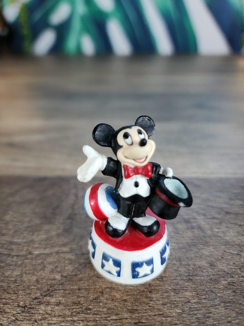 May include: A small figurine of Mickey Mouse dressed in a tuxedo and red bow tie, holding a top hat. He stands on a red, white, and blue circus-style platform with star designs. A red, white, and blue ball is also present.
