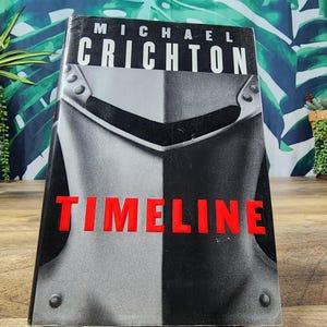May include: A hardcover book titled Timeline by Michael Crichton. The front cover features a black and white illustration of a metallic knight's helmet with rivets. The author's name is printed in white at the top, and the title is in bold red.