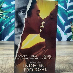 May include: A VHS tape for the movie "Indecent Proposal" featuring Robert Redford, Demi Moore, and Woody Harrelson. The cover art shows a man's face and a couple kissing, with a yellow and orange background.
