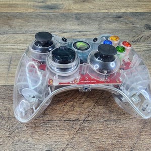May include: Clear plastic video game controller with black joysticks and colored buttons. The controller has a transparent design, revealing the internal components. The controller is on a wooden surface.