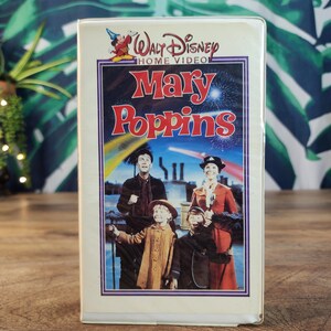 Peut inclure: Une cassette VHS Walt Disney Home Video blanche pour le film Mary Poppins. La couverture présente le titre en rouge, une illustration des personnages principaux et une petite icône de Mickey Mouse dans le coin supérieur gauche sur un ciel bleu.