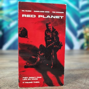 Peut inclure: Une cassette VHS rectangulaire du film Red Planet avec un fond rouge vif. La couverture présente deux personnages en combinaison spatiale, le texte Val Kilmer, Carrie-Anne Moss, Tom Sizemore, Red Planet, et They didn't find life on Mars. It found them.