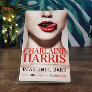 May include: A paperback book titled "Dead Until Dark" by Charlaine Harris. The cover features a close-up of a person's mouth with red lipstick and a drop of liquid. The book is displayed on a clear stand.