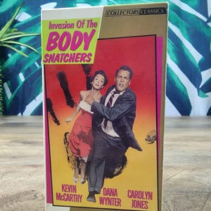 Peut inclure: Une cassette VHS du film Invasion of the Body Snatchers, avec un graphique d'un homme et d'une femme courant sur un fond rouge et jaune. Le texte indique Invasion Of The Body Snatchers, Collectors Classics, Kevin McCarthy, Dana Wynter, Carolyn Jones et Republic Pictures Home Video.