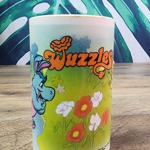 Könnte beinhalten: Ein weißer Keramikbecher mit einer bunten Grafik des Wuzzles-Logos in orangefarbenem Text über einer blauen Zeichentrickfigur und verschiedenen roten und weißen Blumen auf grünem Grund. Der Boden trägt den Text 1985 HASBRO INC. WALT DISNEY.