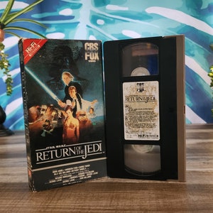 May include: A vintage VHS tape of "Star Wars: Return of the Jedi" with its original box. The box features a movie poster with characters and the film title. The tape is open, revealing the cassette inside.
