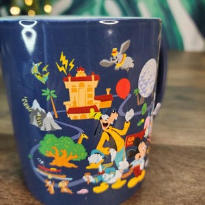 May include: A navy blue ceramic mug featuring colorful illustrations of Disney characters and landmarks. The design includes Goofy, Donald Duck, Mickey Mouse, Dumbo, and a red balloon. The mug has a white handle.