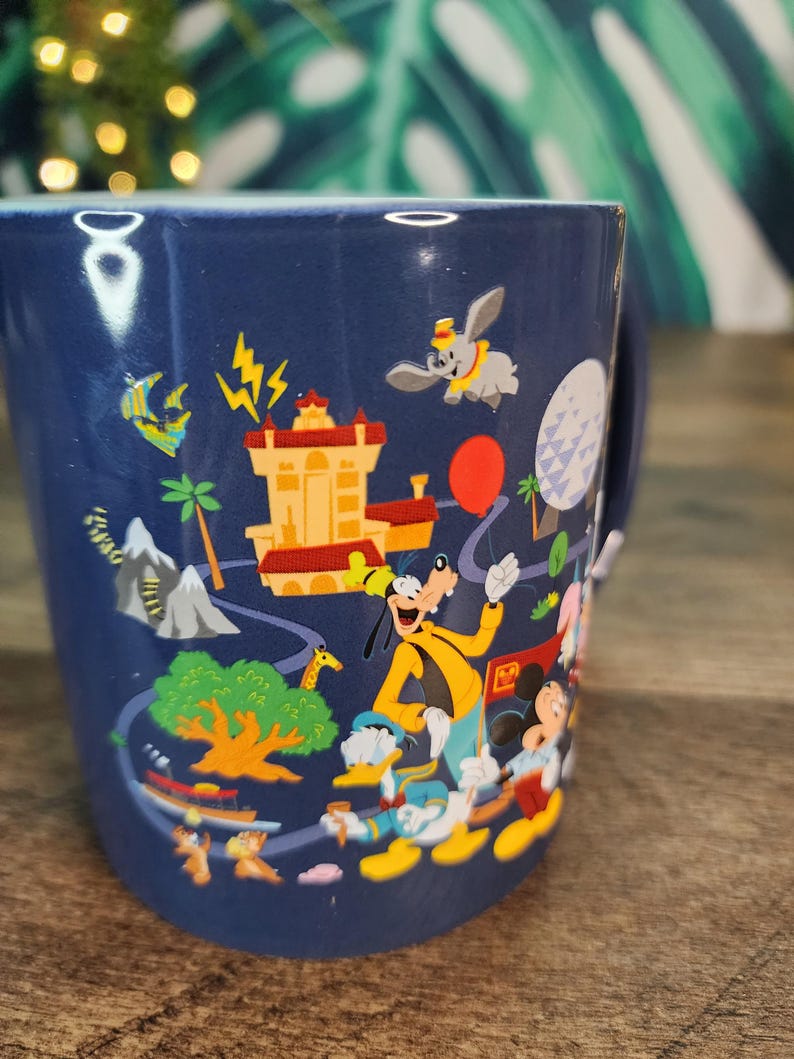 May include: A navy blue ceramic mug featuring colorful illustrations of Disney characters and landmarks. The design includes Goofy, Donald Duck, Mickey Mouse, Dumbo, and a red balloon. The mug has a white handle.
