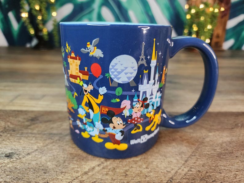 May include: A navy blue ceramic mug featuring a colorful illustration of Disney characters and landmarks. The design includes Mickey Mouse, Minnie Mouse, Goofy, and Dumbo, with the Eiffel Tower and Cinderella's castle. The mug has a large handle.