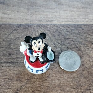 May include: A small Mickey Mouse figurine in a tuxedo and top hat, standing on a red, white, and blue base. A silver coin is next to the figurine. The figurine is approximately 2 inches tall.