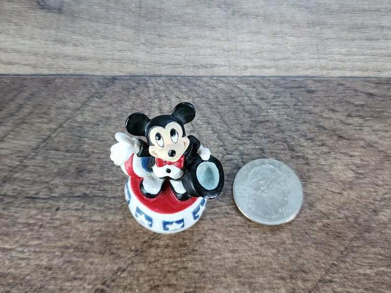 May include: A small Mickey Mouse figurine in a tuxedo and top hat, standing on a red, white, and blue base. A silver coin is next to the figurine. The figurine is approximately 2 inches tall.