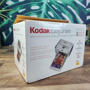 May include: A white cardboard box for a Kodak EasyShare printer dock plus. The box features the Kodak EasyShare logo, text in English, French, and Spanish, and a graphic showing the silver printer dock plus with a printed photograph of a child.