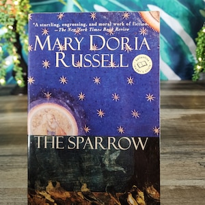 May include: A paperback book titled The Sparrow by Mary Doria Russell. The cover features a dark blue starry sky background with gold stars, a circular illustration of a figure, and a bottom section depicting birds in a dark, earthy landscape.