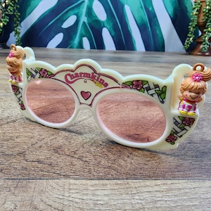 May include: Off-white plastic sunglasses featuring pink-tinted lenses and the text Charmkins printed in red. The frame is decorated with a lattice pattern, small pink flowers, and a central heart. Two small doll figures are attached to the top corners.