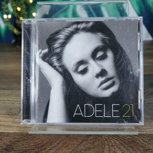 May include: A CD case featuring a black and white image of Adele. The album cover displays the text "ADELE 21" in white and green. The CD case is clear and sits on a clear stand.