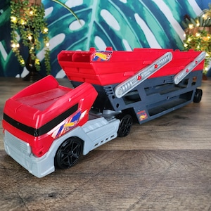 Puede incluir: Un camión de juguete transportador de coches Hot Wheels en color rojo y gris con cabina desmontable y remolque de dos niveles. El vehículo cuenta con ruedas negras, una cabina roja con detalles gráficos en azul y blanco, y un chasis gris.