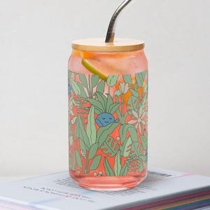May include: A pink glass can with a bamboo lid and a metal straw. The can has a colorful floral pattern with a blue creature inside. The can is sitting on top of a stack of books.