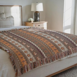 May include: A woven throw blanket with a brown background and a repeating pattern of pumpkins, horses, and geometric shapes in orange, white, and burgundy. The blanket has a fringed edge and is draped over a bed.