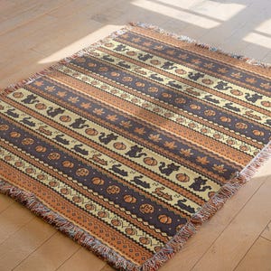 Puede incluir: Una manta rectangular con un patrón repetido de calabazas, hojas y conejos en tonos marrones, naranjas y crema. La manta tiene un borde con flecos y está tejida.