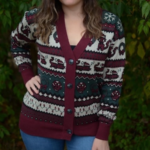 May include: A burgundy cardigan sweater with a festive pattern featuring snowflakes, reindeer, and other holiday motifs. The sweater has a V-neck, button closure, and ribbed cuffs. The background is a natural green.