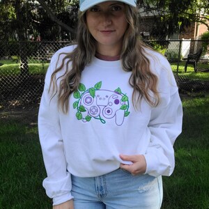 Peut inclure: Un sweat-shirt blanc avec un motif de manette GameCube violet et vert. La manette est entourée de vignes. Le modèle porte un jean bleu et une casquette de baseball bleu clair avec un motif de manette GameCube.