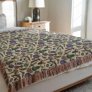 May include: A beige throw blanket with a repeating pattern of fantasy game-themed illustrations, including swords, bows, and potions. The blanket has a fringed edge in shades of brown, orange, and red. The blanket is draped over a bed.