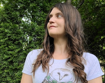 May include: A white t-shirt with a graphic of a barn owl wearing a green and white striped scarf. The owl is perched on a branch.