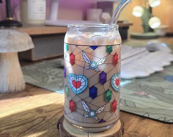 May include: A glass can-shaped cup filled with a light-colored beverage and ice, resting on a wooden coaster. The cup is decorated with a stained-glass design featuring hearts, gems, and winged emblems. A clear glass straw extends from the cup.