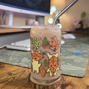 May include: A glass can-shaped cup filled with a light brown beverage and ice, featuring a stainless steel straw. The cup is decorated with pixelated autumn-themed graphics of leaves and creatures. It rests on a wooden coaster.