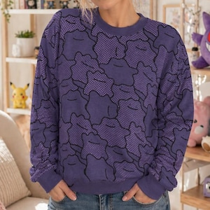 May include: A purple sweatshirt with a black pattern is worn with blue jeans. The sweatshirt has a crew neck and long sleeves. The jeans are distressed with rips at the knees. The background includes shelves with figurines and a sign that says "Good Day Good Coffee Good Game."