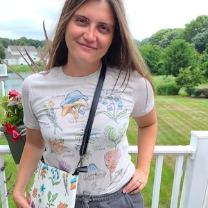 May include: A woman wearing a white t-shirt with a mushroom and flower print and grey sweatpants with a green and black video game controller graphic. She is carrying a white crossbody bag with a floral print.