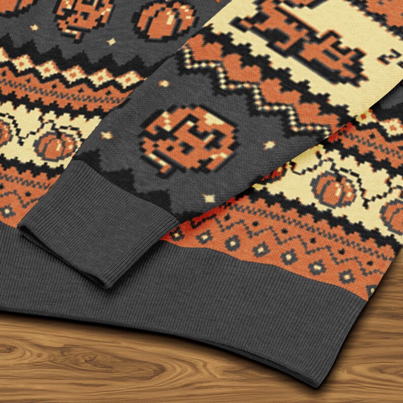 Puede incluir: Primer plano de un su&eacute;ter gris de Halloween con calabazas pixeladas de color naranja y crema y patrones geom&eacute;tricos. El su&eacute;ter tiene un pu&ntilde;o gris acanalado y cuello redondo. El dise&ntilde;o recuerda a los gr&aacute;ficos de videojuegos retro.