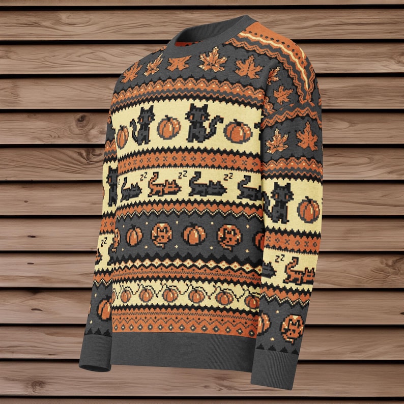 Puede incluir: Un su&eacute;ter de Halloween gris, naranja y crema con gatos negros pixelados, calabazas y hojas de oto&ntilde;o. El su&eacute;ter tiene cuello, pu&ntilde;os y dobladillo de canal&eacute;. El dise&ntilde;o se repite en la parte delantera y las mangas.
