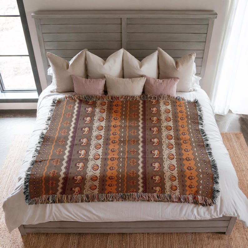 May include: A brown and orange patterned throw blanket with fringe. The blanket features repeating patterns of elephants, circles, and zigzags. The throw is displayed on a bed with pillows.