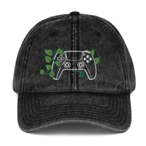 以下が含まれることがあります： 緑のつるが巻き付いたビデオゲームコントローラーの刺繍デザインが施された、黒いヴィンテージデニムのベースボールキャップです。